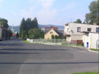 Kozlovice: Pohled na cestu v obci, vpravo autobusová zastávka, v pozadí místní kostel